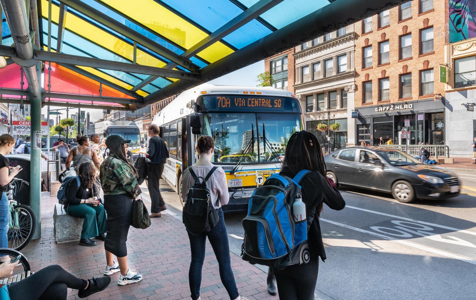 70A bus pulling up to the Central Square stop, with backpack-wearing riders waiting