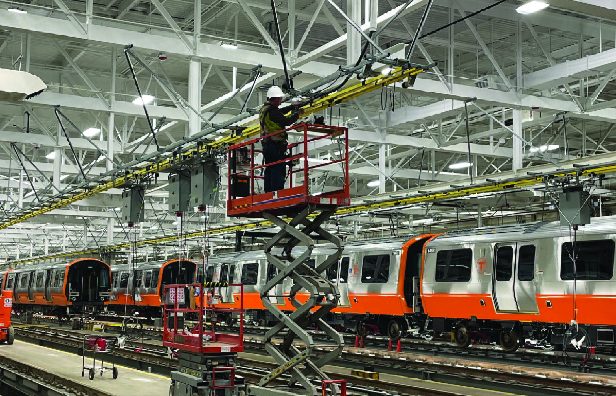 A crew member in a lift works on a rail near the ceiling inside the vehicle maintenance facility